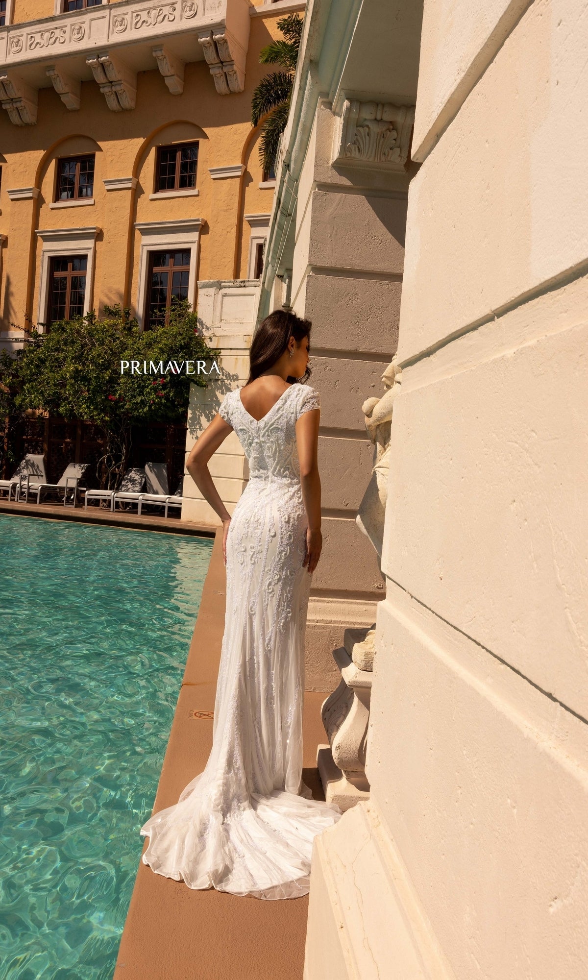 A woman in the Long Wedding Dress 11121 by Primavera, featuring an ivory hue and v-neckline, stands beside a pool near a beige building, facing away from the camera.